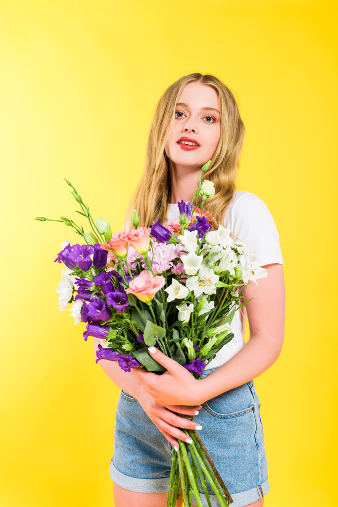 attraktives blondes Mädchen mit Blumen blickt in die Kamera auf gelb - Foto, Bild