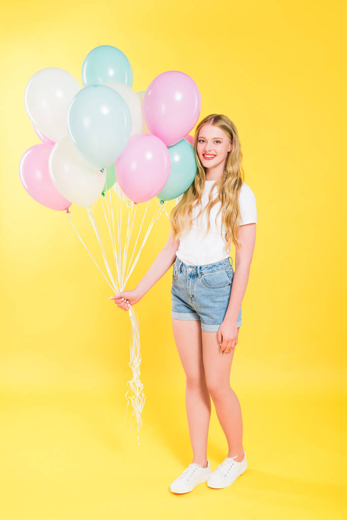 όμορφο κορίτσι με μπαλόνια κοιτάζοντας την κάμερα και χαμογελά στο κίτρινο - Φωτογραφία, εικόνα