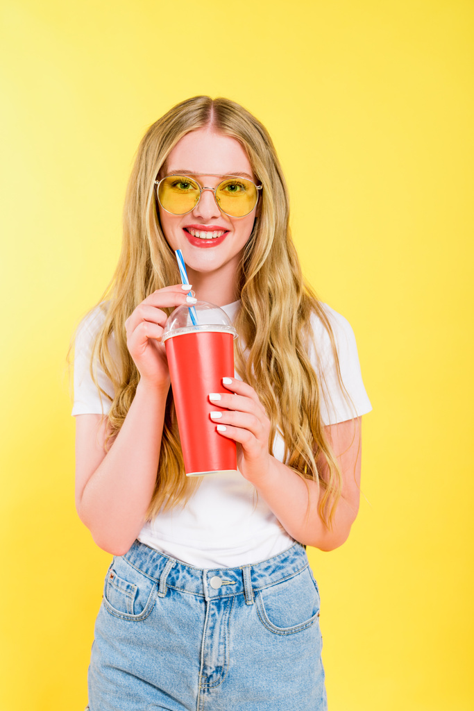 mooi glimlachend meisje in zonnebril met drankje in wegwerp beker geïsoleerd op geel  - Foto, afbeelding