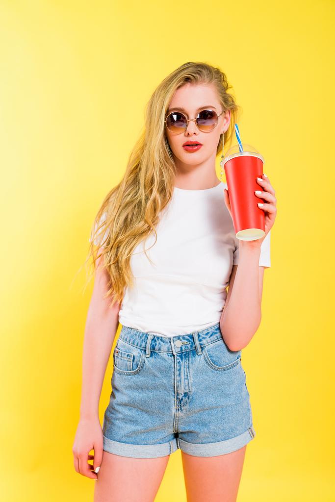 schönes Mädchen mit Sonnenbrille und Getränk im Einwegbecher auf gelb  - Foto, Bild
