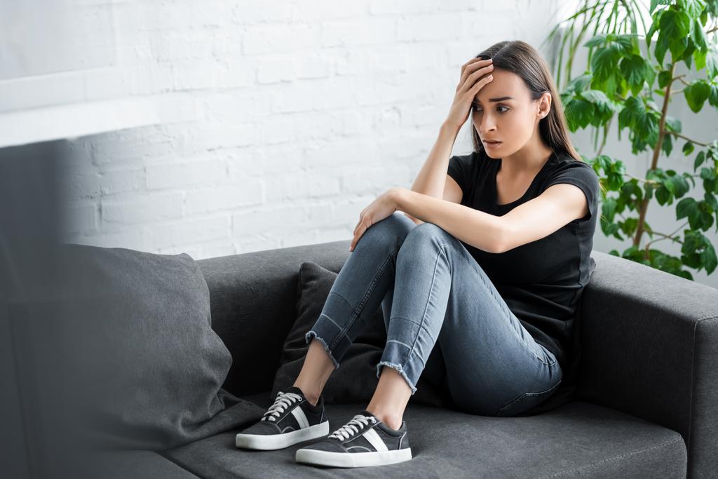 bonita jovem mulher que sofre de depressão enquanto sentado no sofá em casa e segurando a mão perto da cabeça
 - Foto, Imagem
