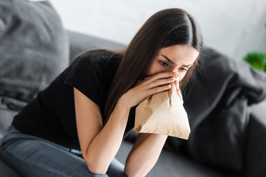 νεαρή γυναίκα αναπνέει σε χάρτινη σακούλα ενώ υποφέρει από κρίση πανικού στο σπίτι - Φωτογραφία, εικόνα