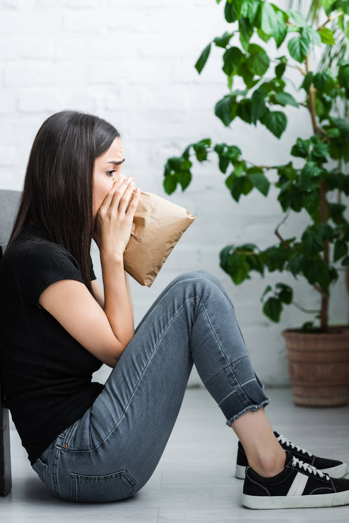side view of young woman sitting on floor and breathing into paper bag while suffering from panic attack - Photo, Image
