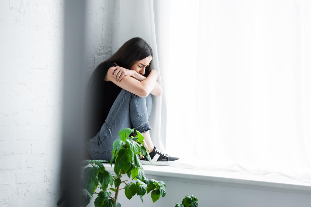 depressed young woman crying while sitting on window sill and hiding face in crossed arms - Photo, Image