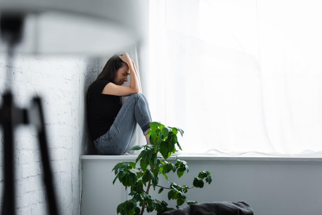 επιλεκτική εστίαση της καταθλιπτικής νεαρής γυναίκας που κλαίει ενώ κάθεται στο περβάζι του παραθύρου και κρατώντας τα χέρια στο κεφάλι - Φωτογραφία, εικόνα