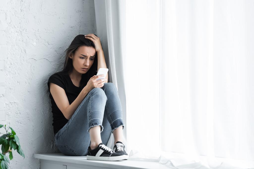 mujer joven deprimida sentada en el alféizar de la ventana en casa y usando teléfono inteligente
 - Foto, Imagen