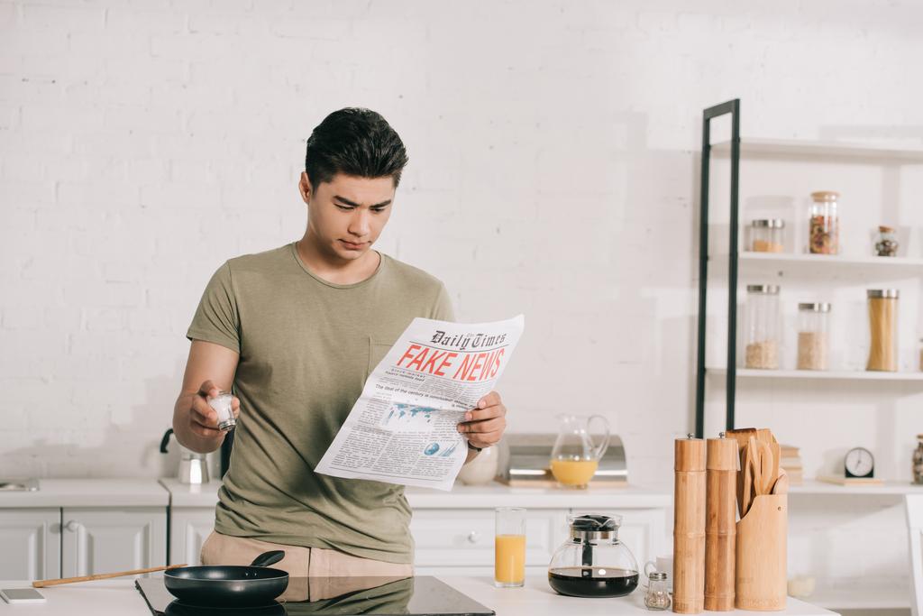 красивый азиатский мужчина готовит завтрак во время чтения фейковых новостей газеты на кухне
 - Фото, изображение