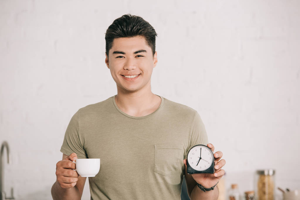 カメラに微笑みながらコーヒーカップと目覚まし時計を見せる陽気なアジア人男性 - 写真・画像