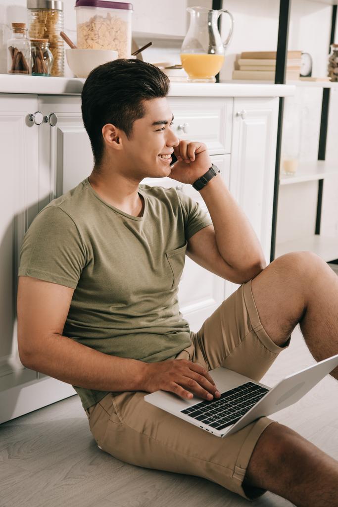 mosolygós ázsiai ember beszél okostelefonon és használ laptop ülve a padlón a konyhában - Fotó, kép