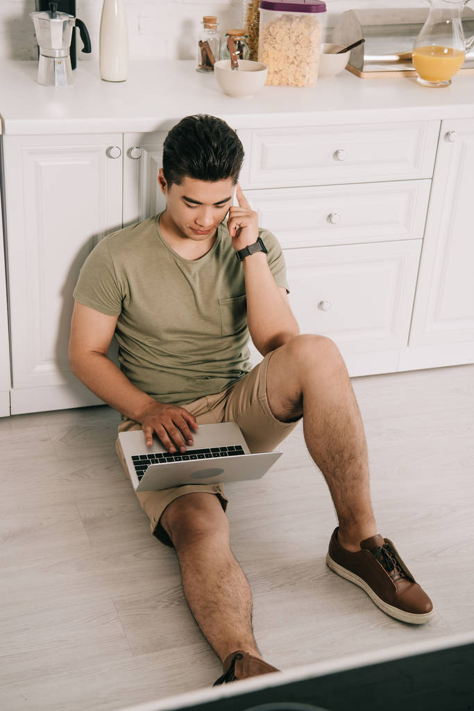 high angle view of young asian man talking on smartphone and using laptop while sitting on floor in kitchen - Photo, Image