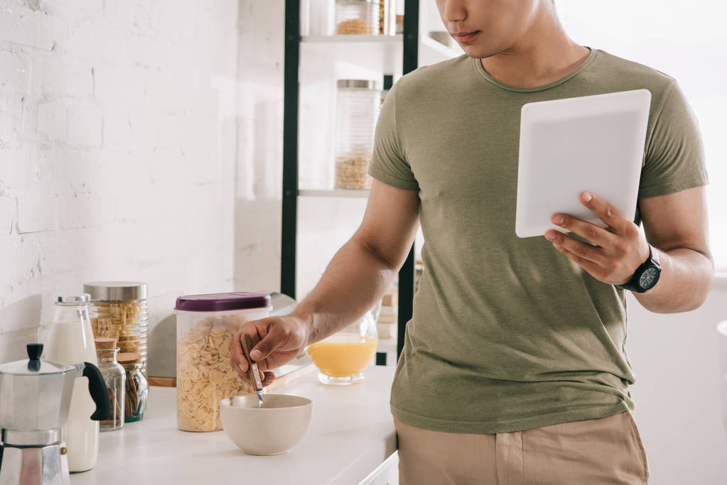 Teilbild eines jungen Mannes, der Flocken in Schüssel mixt, während er digitales Tablet in der Küche hält - Foto, Bild