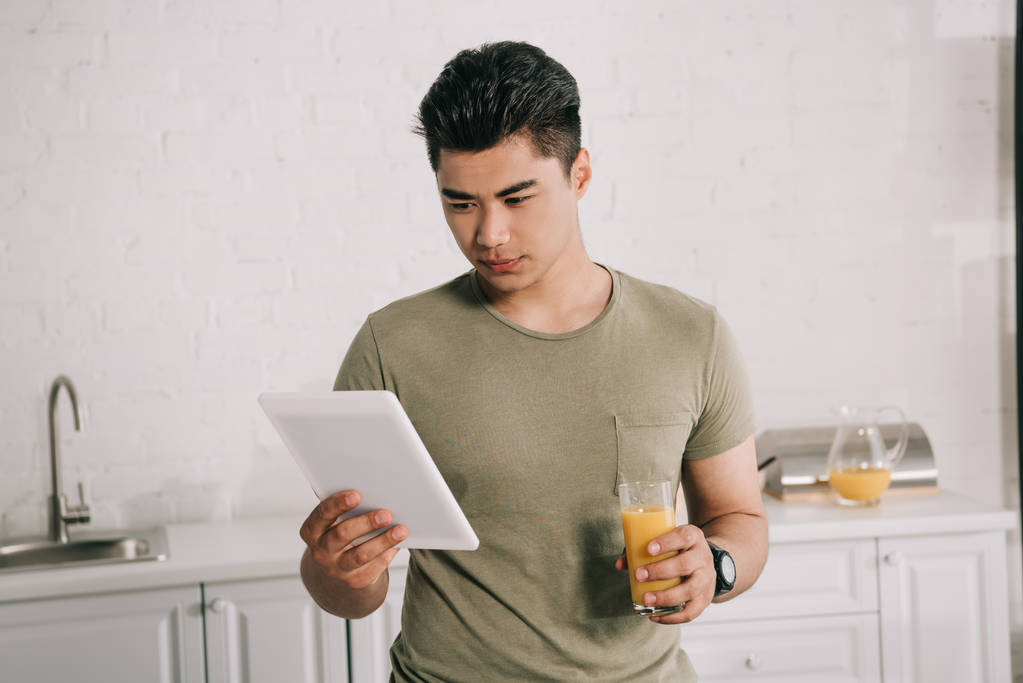 giovane uomo asiatico utilizzando tablet digitale mentre in piedi in cucina con vetro di succo d'arancia
 - Foto, immagini