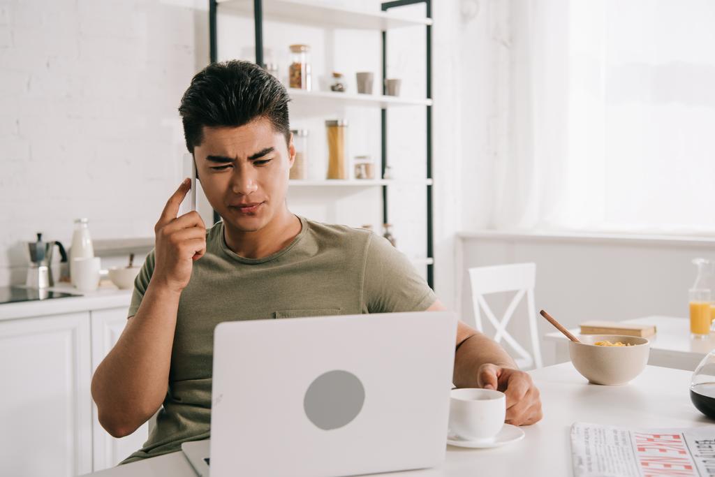 серйозний азіатський чоловік говорить на смартфоні, сидячи за кухонним столом і використовуючи ноутбук
 - Фото, зображення