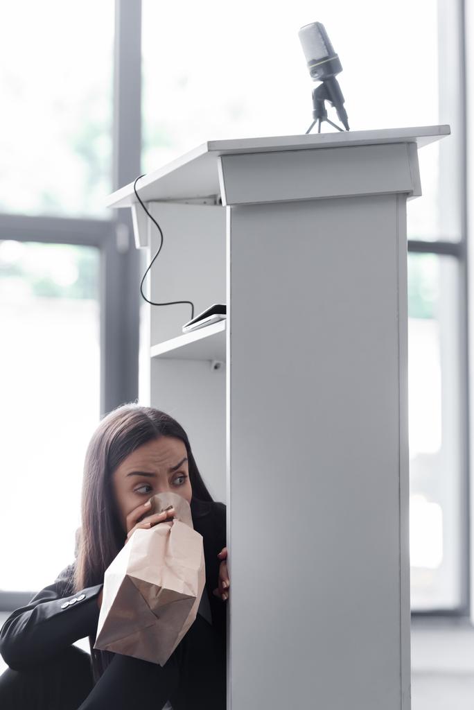 испуганный лектор дышит в бумажный пакет, сидя на полу в конференц-зале и страдая от панической атаки
 - Фото, изображение