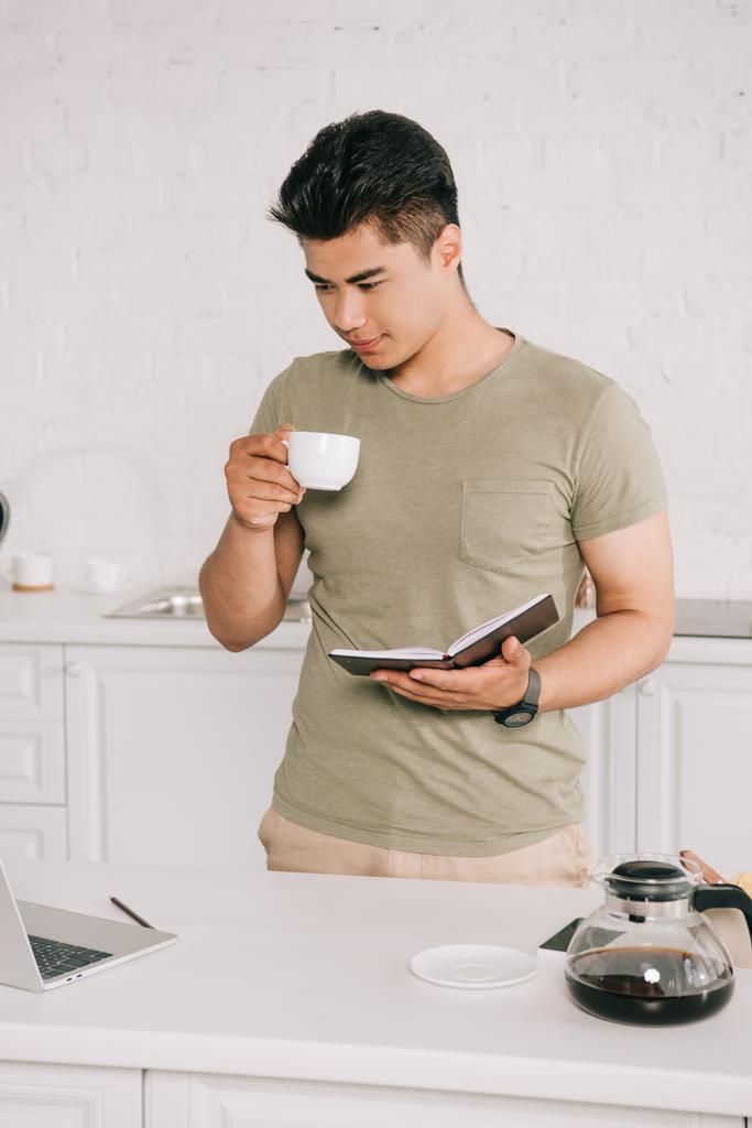 молодий азіатський чоловік п'є каву, дивлячись на ноутбук і тримаючи блокнот
 - Фото, зображення