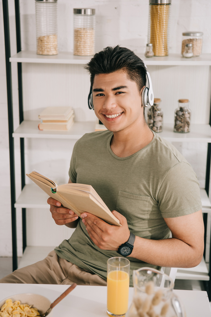 Veselý asijský muž ve sluchátkách s úsměvem na kameře, když sedí u kuchyňského stolu a drží knihu - Fotografie, Obrázek