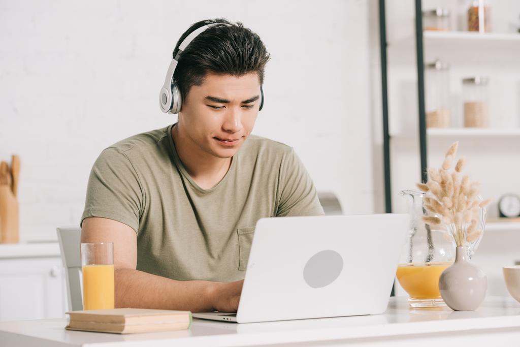 concentrado asiático homem em fones de ouvido usando laptop enquanto sentado na mesa da cozinha
 - Foto, Imagem