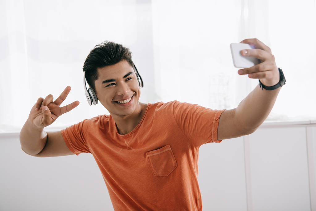 szczęśliwy Asian Mężczyzna biorąc selfie pokazując gest zwycięstwa i słuchania muzyki w słuchawkach - Zdjęcie, obraz