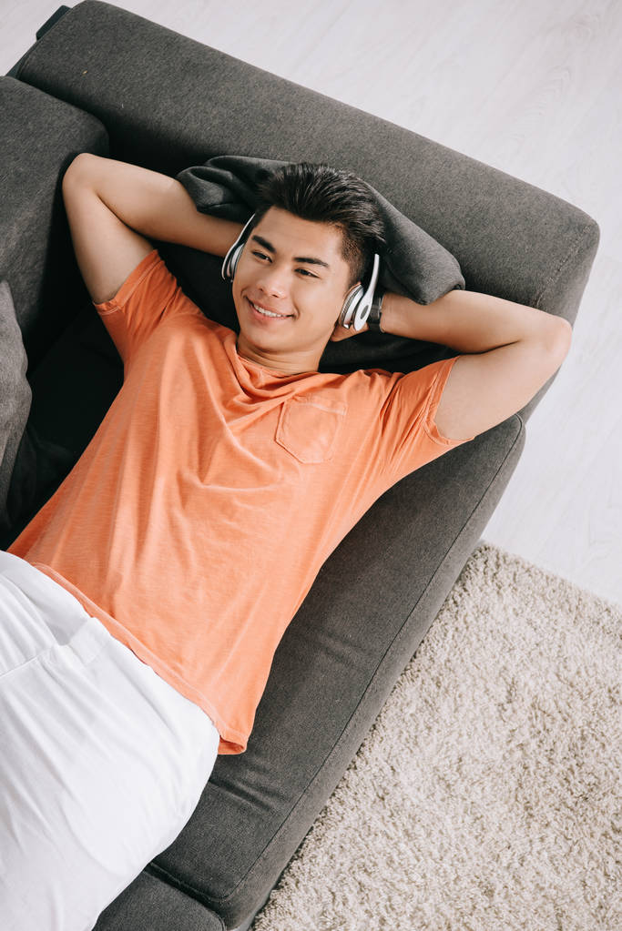 Draufsicht eines lächelnden asiatischen Mannes, der auf dem Sofa liegt und über Kopfhörer Musik hört - Foto, Bild