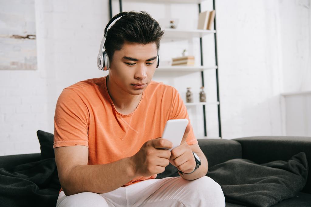 στοχαστικός Ασιάτης άνθρωπος ακούγοντας μουσική στα ακουστικά και χρησιμοποιώντας smartphone, ενώ κάθεται στον καναπέ στο σπίτι - Φωτογραφία, εικόνα