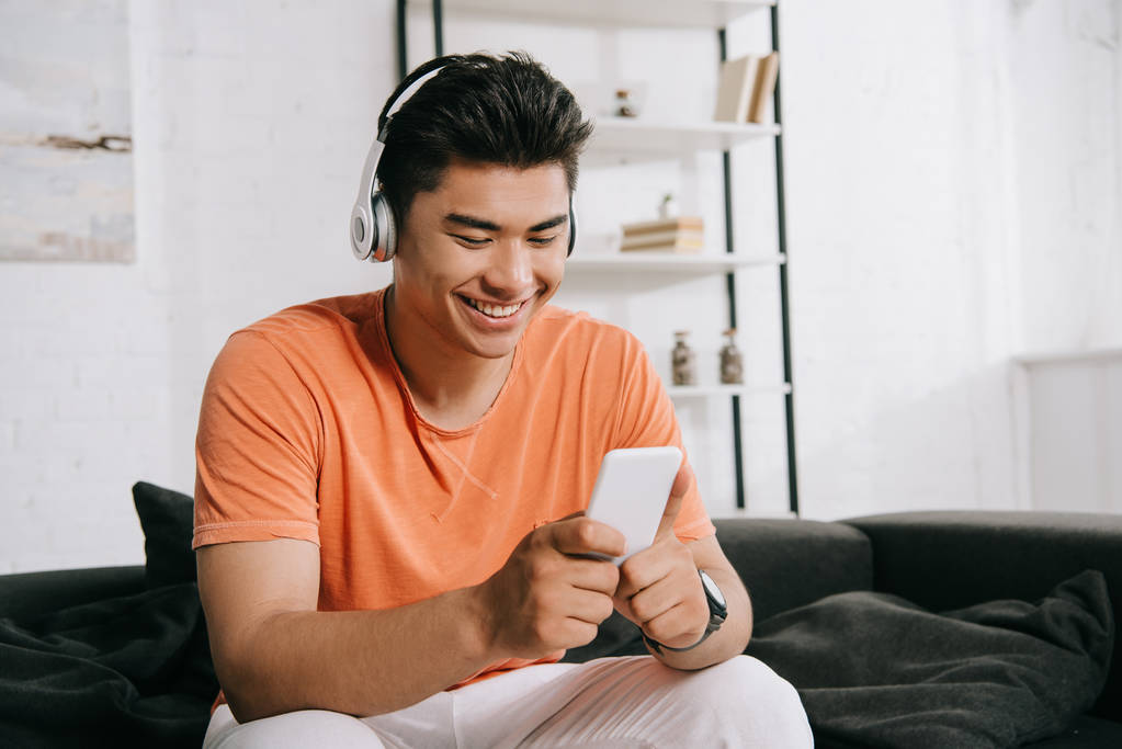 allegro uomo asiatico utilizzando smartphone e ascoltare musica in cuffia mentre seduto sul divano a casa
 - Foto, immagini