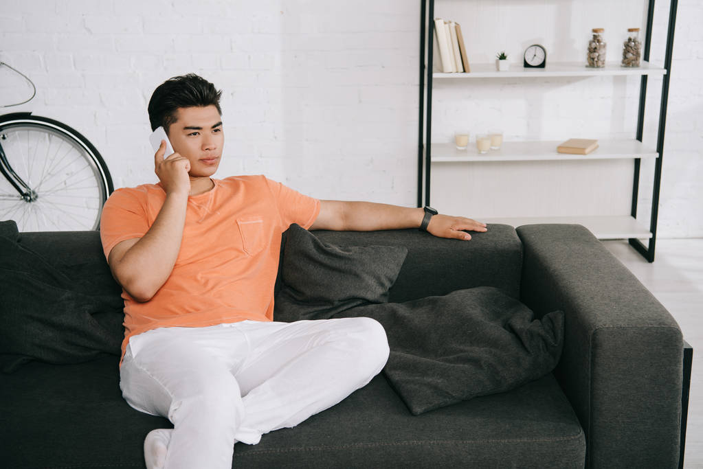 serious asian man talking on smartphone and looking away while sitting on sofa at home - Photo, Image