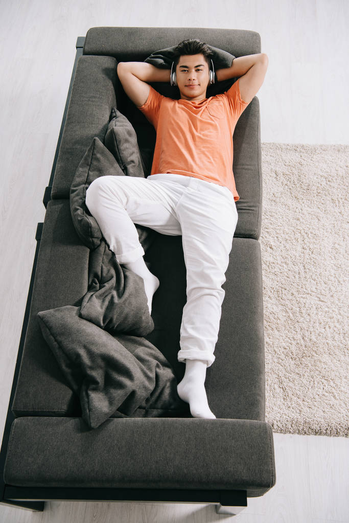 ylhäältä näkymä nuori aasialainen mies kuuntelee musiikkia kuulokkeet kun makaa sohvalla kotona
 - Valokuva, kuva