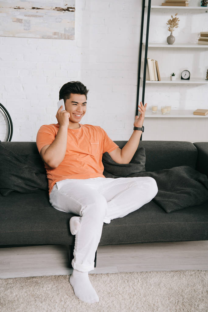 cheerful asian man gesturing while sitting on couch and talking on smartphone - Photo, Image
