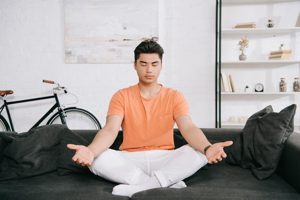 joven asiático hombre meditando mientras sentado en loto pose en sofá en casa
 - Foto, imagen