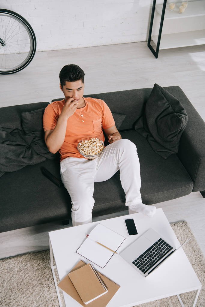 високий кут зору молодого азіатського чоловіка, який їсть попкорн, сидячи на дивані біля столу з ноутбуком і блокнотами
 - Фото, зображення