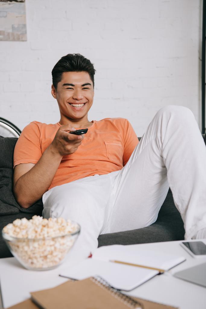 wesoły Asian Mężczyzna oglądanie TV podczas siedzący na tapczan blisko stół z Puchar z popcorn - Zdjęcie, obraz