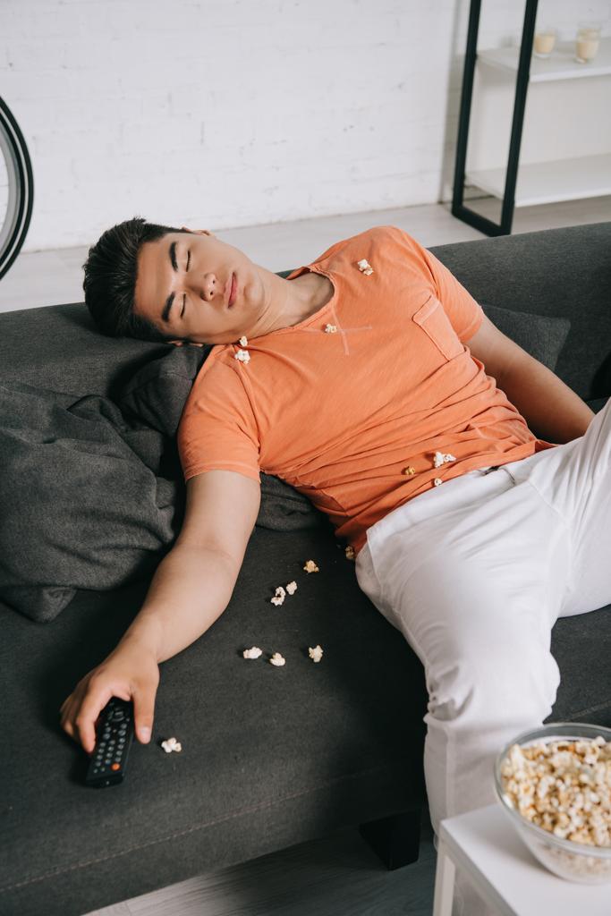agotado asiático hombre durmiendo en sofá cerca de mesa con bowl de palomitas de maíz
 - Foto, imagen