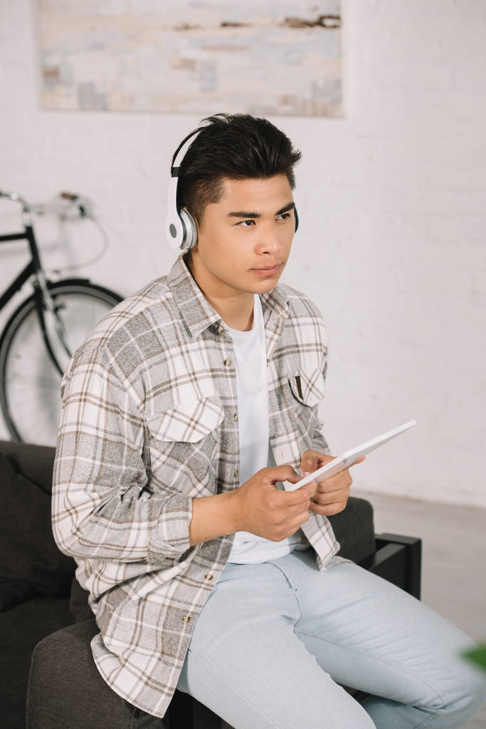 cher asiatique homme regardant loin tout en écoutant de la musique dans casque et tenant tablette numérique
 - Photo, image