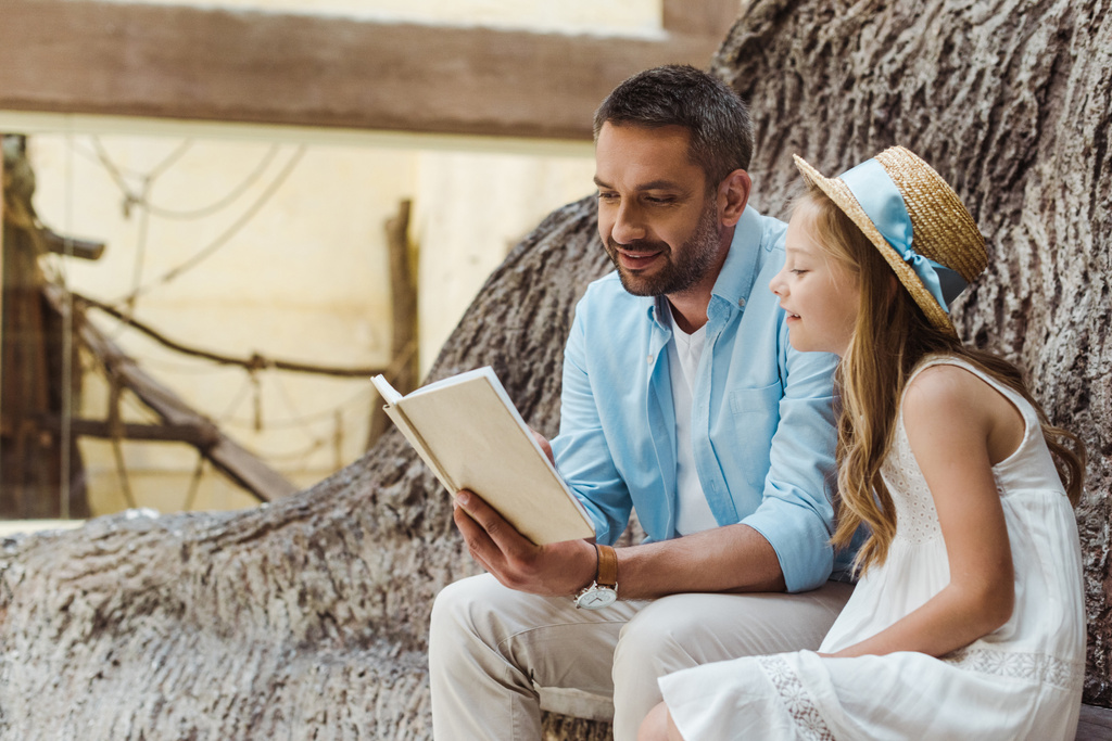 cheerful father reading book near cute daughter in straw hat near tree  - Photo, Image