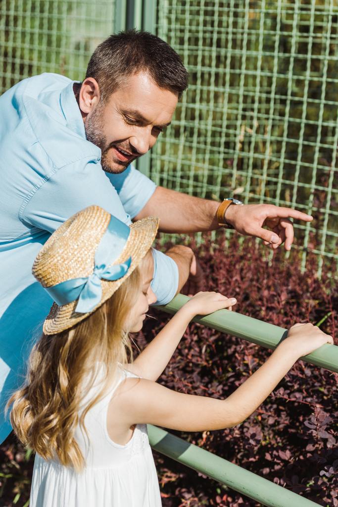 przystojny mężczyzna wskazując palcem w klatce stojąc z cute córki w zoo  - Zdjęcie, obraz