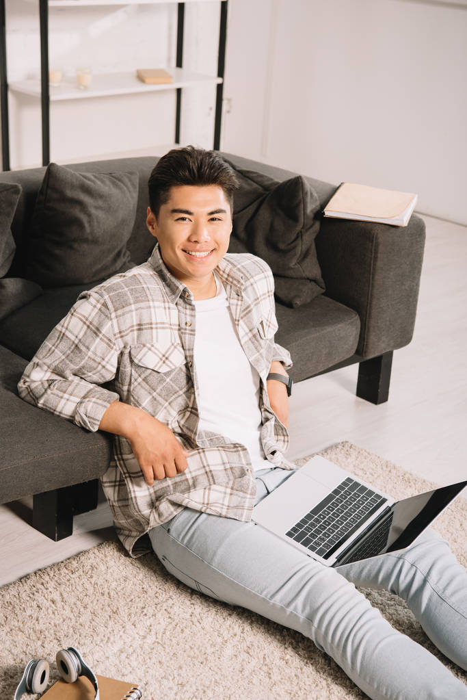 vrolijke Aziatische man glimlachend op camera terwijl zittend op de vloer met laptop - Foto, afbeelding