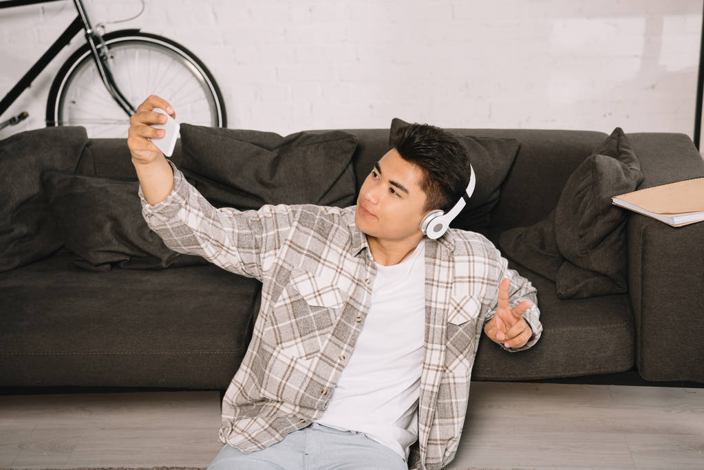 przystojny azjatycki mężczyzna w słuchawkach siedzi na podłodze i biorąc selfie ze smartfonem - Zdjęcie, obraz