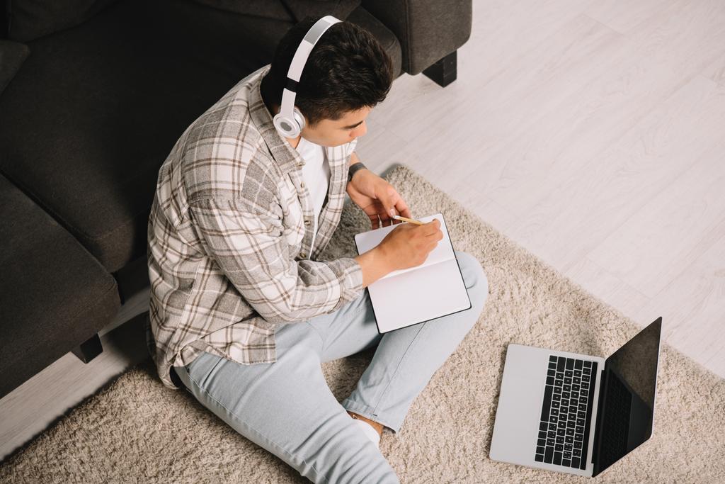 Overhead-Ansicht des jungen Mannes mit Kopfhörer sitzt auf dem Boden in der Nähe von Laptop und schreibt in Notizbuch - Foto, Bild