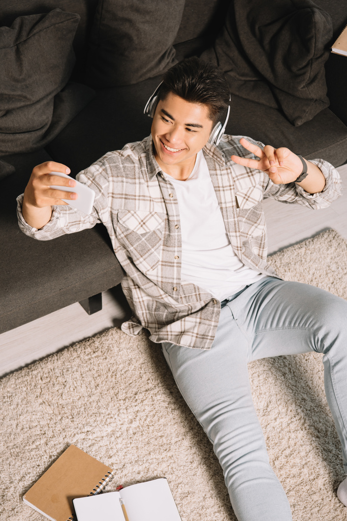 arriba vista de alegre asiático hombre en auriculares tomando selfie mientras sentado en piso
 - Foto, Imagen