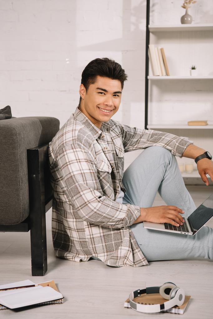 cheerful asian man smiling at camera while sitting on floor at home and using laptop - Photo, Image