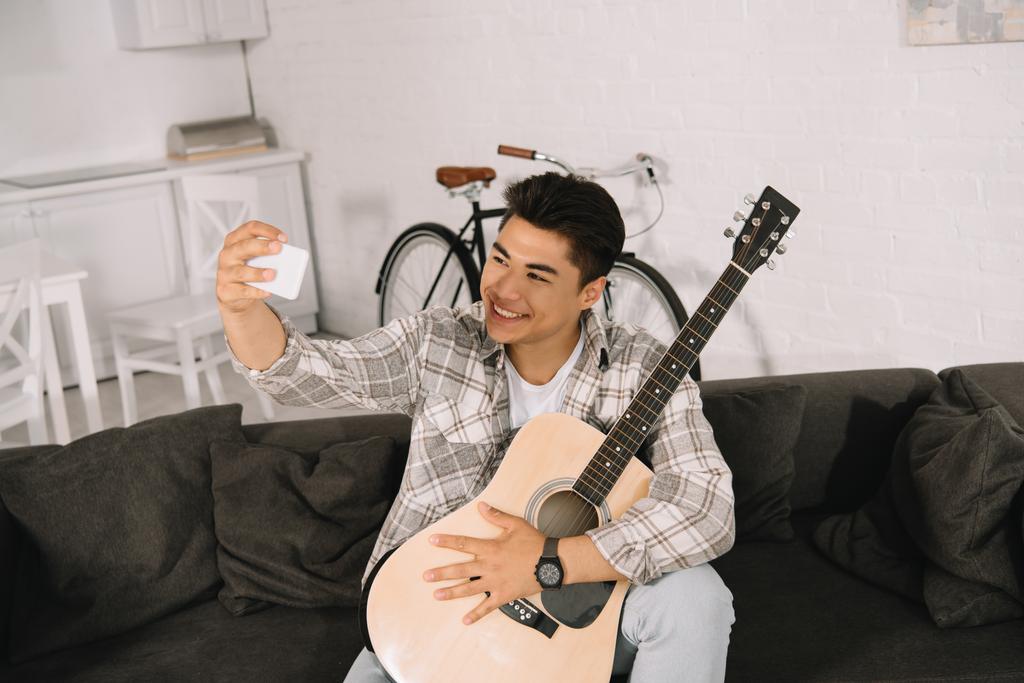 χαρούμενος Ασιάτης/ισσα άνθρωπος λήψη selfie με smartphone, ενώ κάθονται στον καναπέ με ακουστική κιθάρα - Φωτογραφία, εικόνα