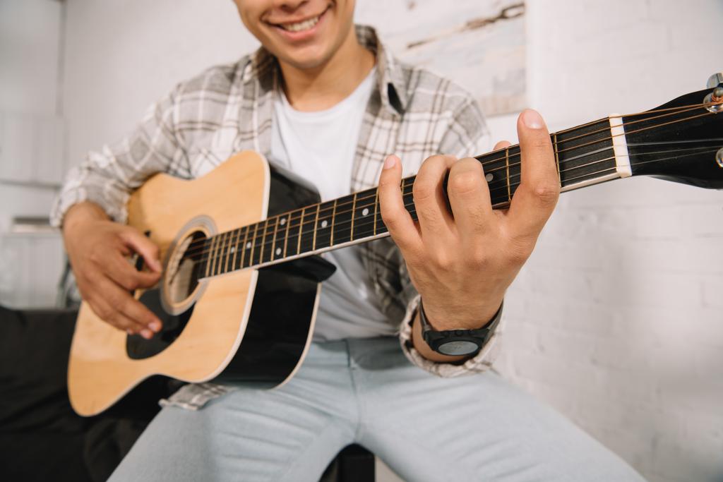 частичный вид улыбающегося молодого человека, играющего на акустической гитаре дома
 - Фото, изображение