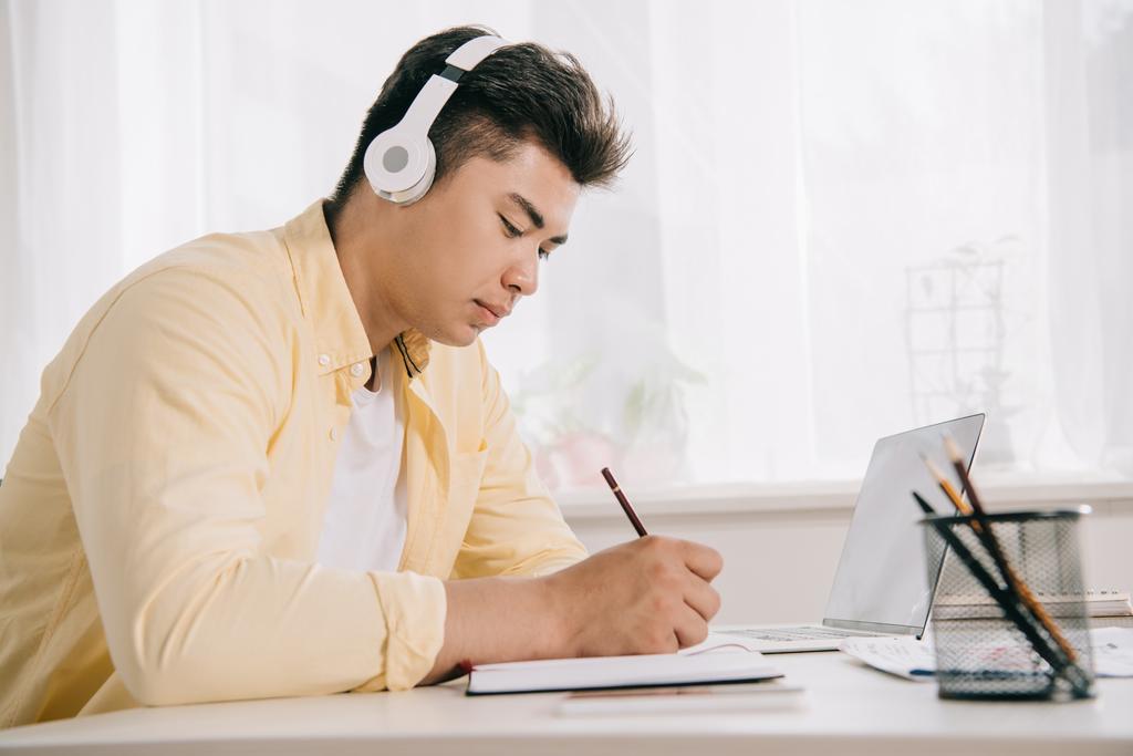 young, concentrated asian man in headphones writing in notebook white sitting at desk - Photo, Image