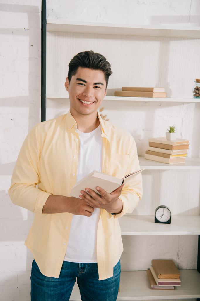 junger, fröhlicher asiatischer Mann mit Buch in der Hand, während er neben dem Regal steht und in die Kamera lächelt - Foto, Bild