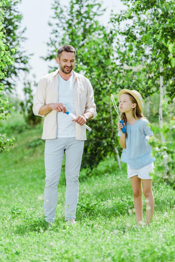 selective focus of cheerful father standing near daughter blowing soap bubbles near trees - Photo, Image