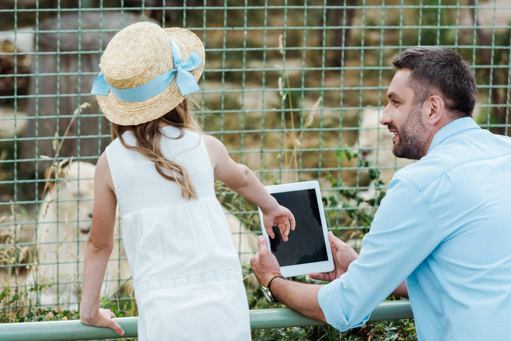 dziecko w słomy kapelusz wskazując w pobliżu cyfrowego tabletu z pustym ekranem i ojcem w zoo  - Zdjęcie, obraz