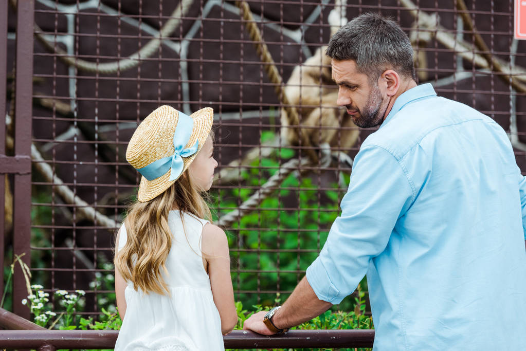 παιδί σε ψάθινο καπέλο κοιτάζοντας αναστατωμένος πατέρας κοντά σε κλουβί με άγριο ζώο στο ζωολογικό κήπο  - Φωτογραφία, εικόνα