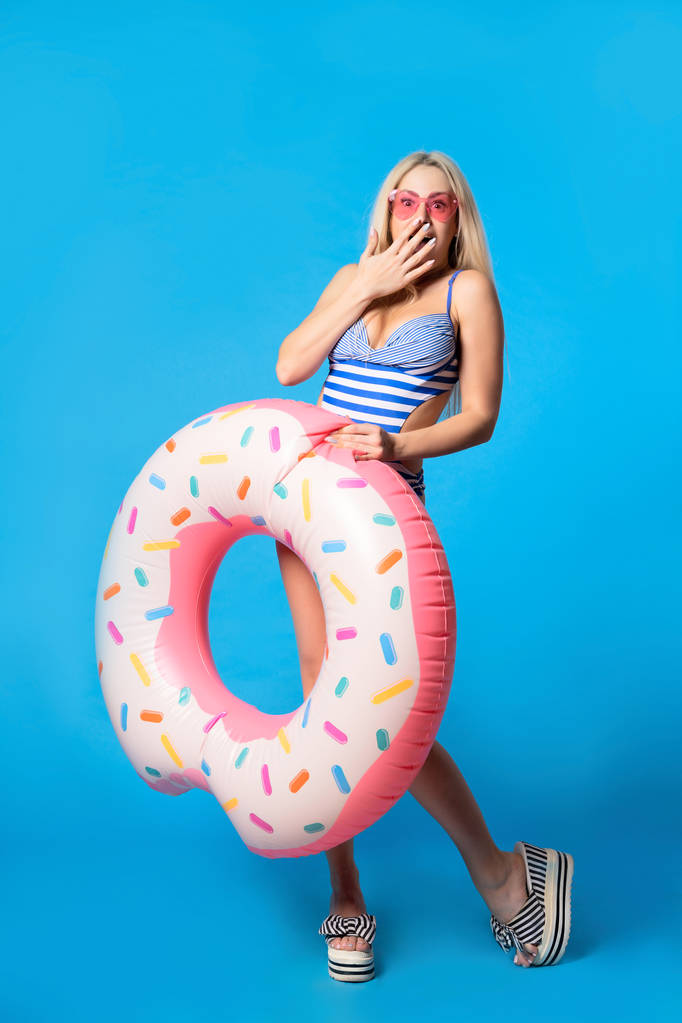 Pełna długość Zdjęcia zaskoczona Kobieta w stroju kąpielowym z nadmuchiwanym pączka na pustym niebieskim tle - Zdjęcie, obraz