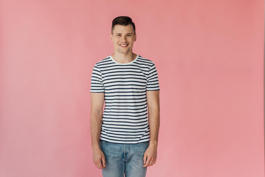 Vorderseite eines lächelnden, gut aussehenden jungen Mannes in gestreiftem T-Shirt, der isoliert auf rosa Kamera blickt - Foto, Bild