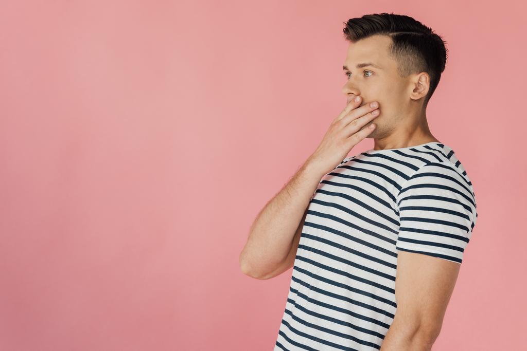 σοκαρισμένος νέος άνδρας με ριγέ t-shirt κάλυψης στόμα με το χέρι απομονωθεί σε ροζ - Φωτογραφία, εικόνα